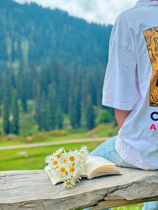 A person in a white graphic t-shirt sits on a wooden bench with an open book and a bouquet of daisies, overlooking a lush green forested mountain valley.