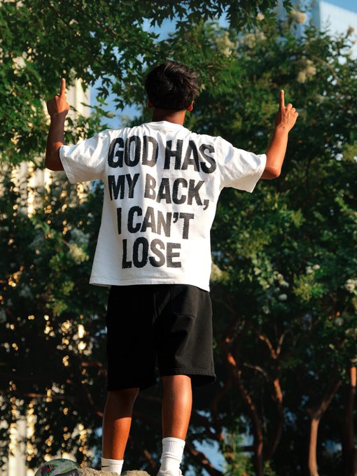 A person standing with their back to the camera, wearing a white t-shirt that reads 'GOD HAS MY BACK, I CAN'T LOSE' in bold black letters, with both hands raised.