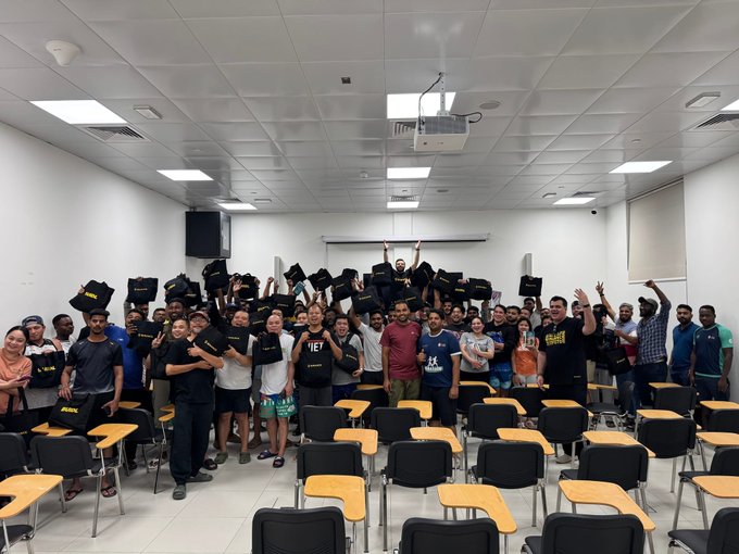 The image shows a group of people standing together in an auditorium or classroom setting with their arms raised high in celebration. They are wearing black t-shirts that say " Crypto is for everyone!". The room has rows of chairs, some of which have backpacks on them, indicating that the event might be related to education or learning about cryptocurrency. The group appears to be engaged and excited about the subject matter being discussed at the workshop they attended.