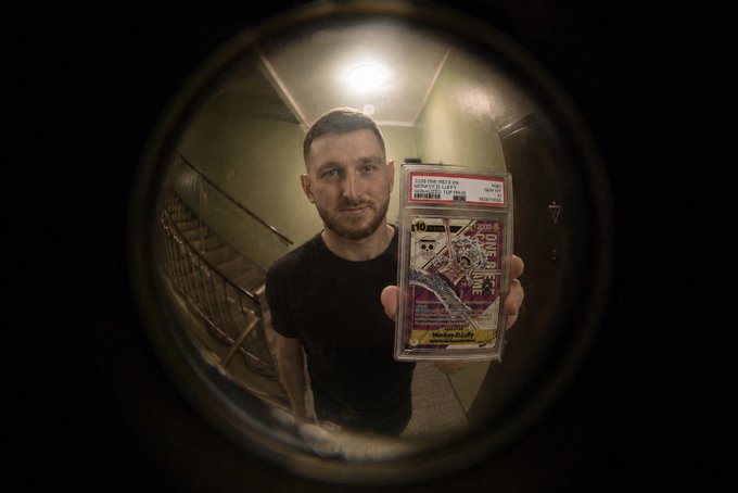 A man holds up a graded 2006 One Piece Monkey D. Luffy "Top Prize" card, as seen through a fisheye peephole, perfectly illustrating the "POV" scenario of the accompanying post.