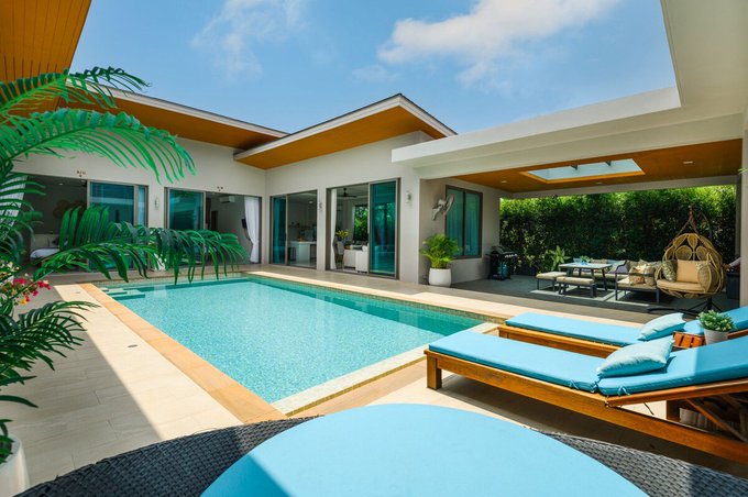 An outdoor patio area featuring a small rectangular swimming pool, lounge chairs with turquoise cushions, and a hanging egg chair beside a modern house.