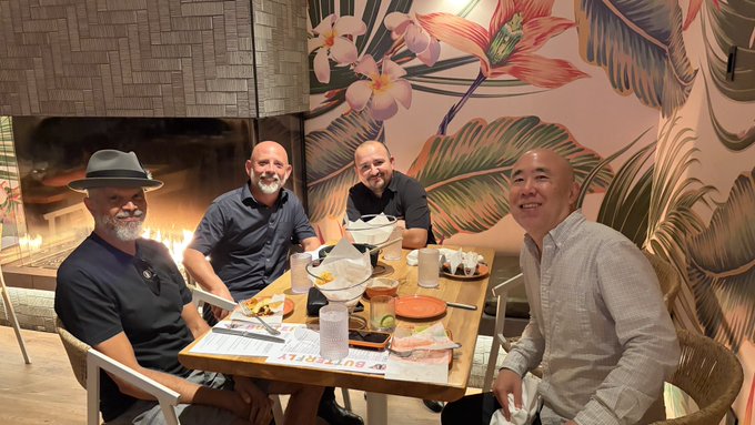 Four men pose around a restaurant table set with food and drinks, seated in front of a tropical-themed mural and a fireplace.
