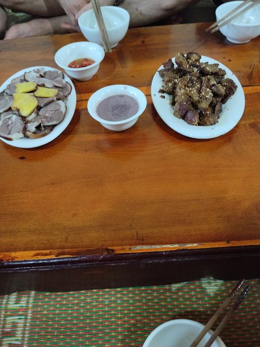A wooden dining table set with bowls and plates of food, including sliced meat with yellow slices, a red sauce, a purple dip, and a plate of sesame-coated fried meat, with chopsticks and white bowls around the edge.