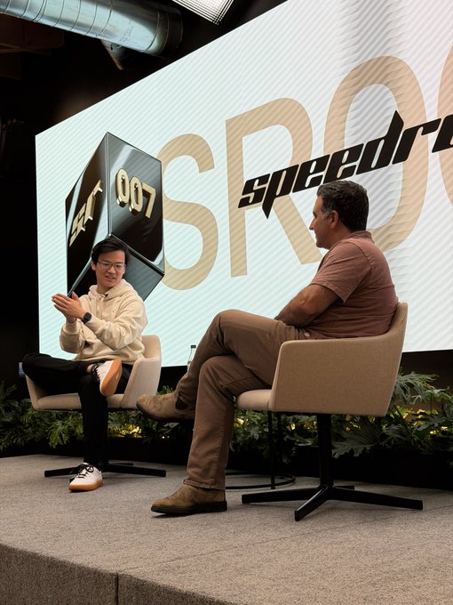 A photo of two men seated on stage in armchairs at what appears to be a 'speedrun' tech event, with one man in a beige hoodie gesturing while speaking to another man in a brown shirt.