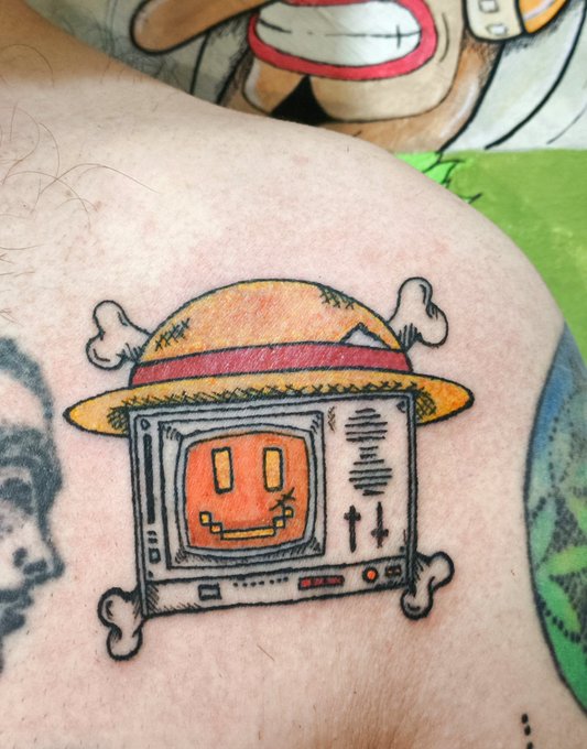A tattoo on skin depicts a cartoonish television character wearing a straw hat, featuring an orange smiling face on the screen and cross-bones decorating the TV casing.