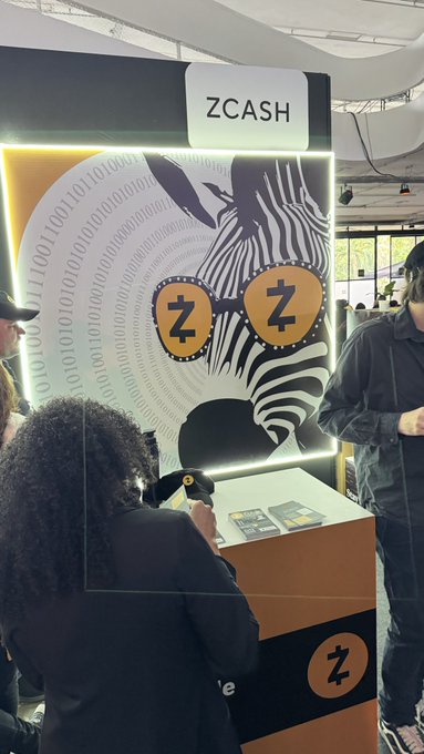 The image features a person wearing a black jacket standing next to a booth with a large poster of a zcash logo behind them. The person appears to be taking a picture or recording something at the booth using their phone, possibly related to the zcash project.