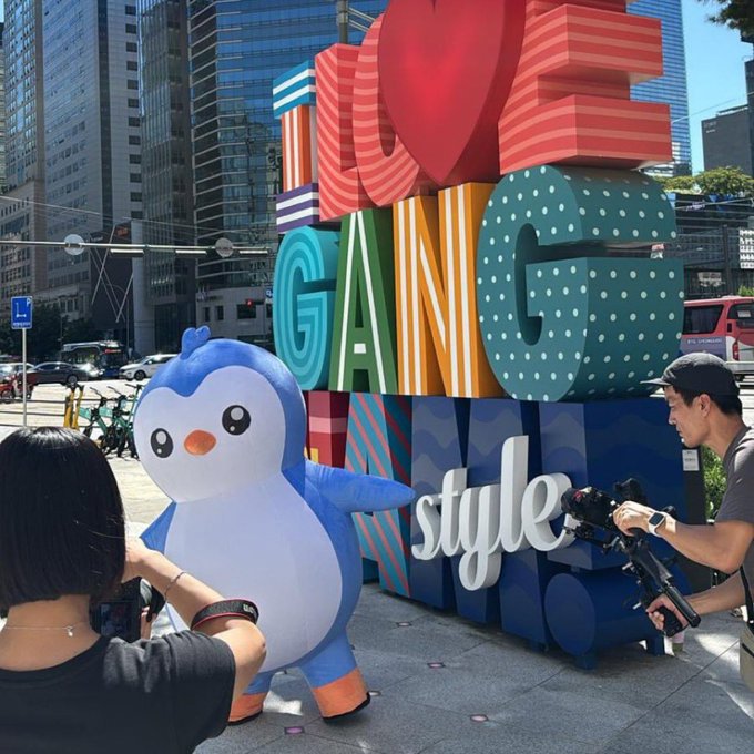 Two people photograph a large inflatable blue and white penguin mascot posing in front of a colorful block-letter sculpture reading 'LOVE GANG style' on a city street.