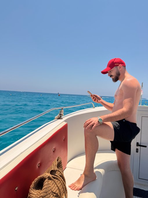 A shirtless man in a red cap and sunglasses sits on a boat, looking down at his phone against a backdrop of blue ocean and sky, sharing a relaxed "Gm friends" moment from his day out at sea.