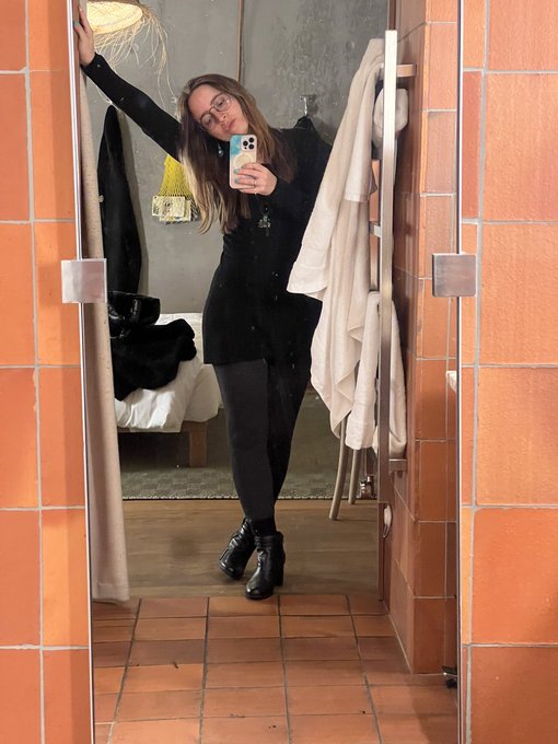 A mirror selfie of a woman wearing glasses, a long black dress, black tights, and black boots, standing in a bathroom with terracotta tiled walls and floors.
