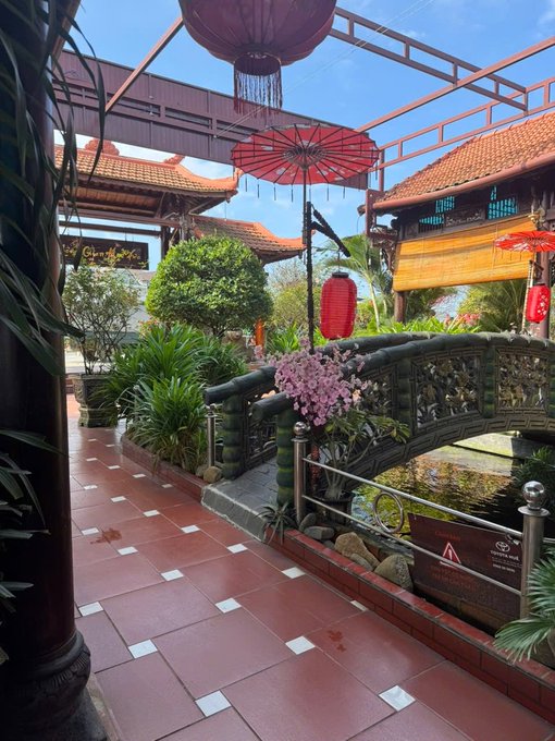 An ornate Asian-style garden walkway with red tiled floors, decorative red lanterns and umbrellas, a small bridge, and traditional architecture.