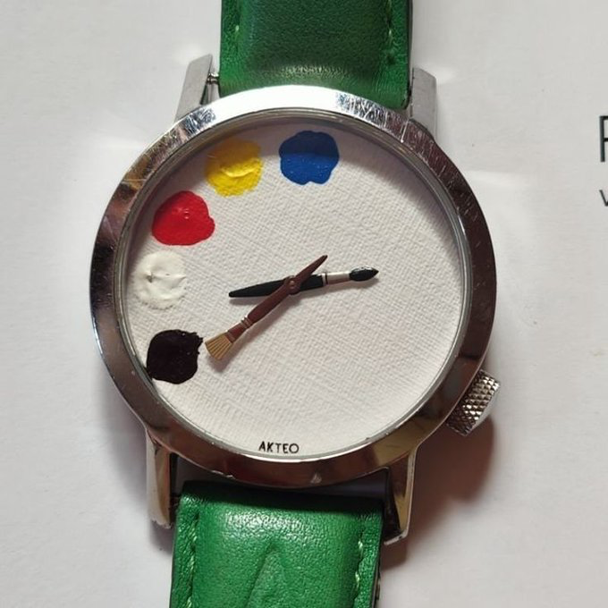 An Akteo wristwatch with a green leather strap and a white canvas dial designed to look like a painter's palette, featuring paint blobs and a paintbrush as the watch hands.