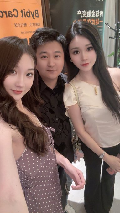 Two young women take a selfie with a man in a black patterned shirt at what appears to be a restaurant, with a 'Bydji Garden' sign visible in the background.
