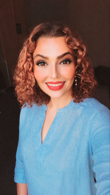 A selfie of a smiling woman with curly auburn hair, dramatic eye makeup, red lipstick, and a blue button-up top in a dimly lit room.