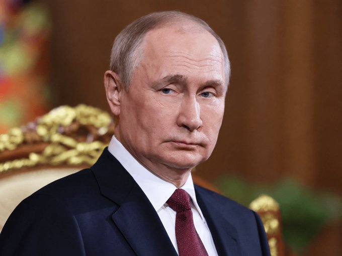 A formal portrait of Russian President Vladimir Putin wearing a dark suit and burgundy tie, seated in an ornate room with a serious expression.