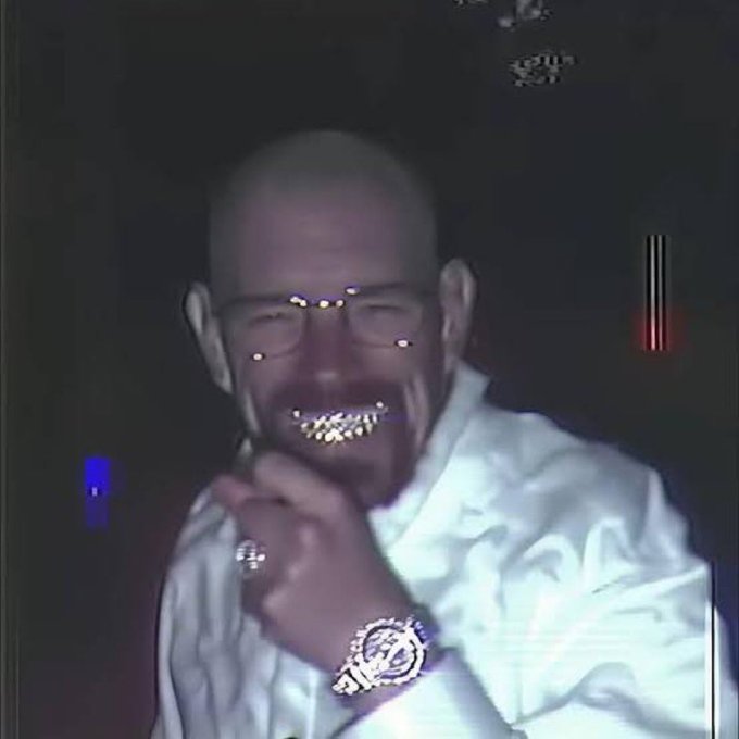 A close-up photo shows a bald man with a beard and glasses, wearing a white shirt and a watch, smiling and holding his hand near his face, with visible diamond-encrusted grillz.