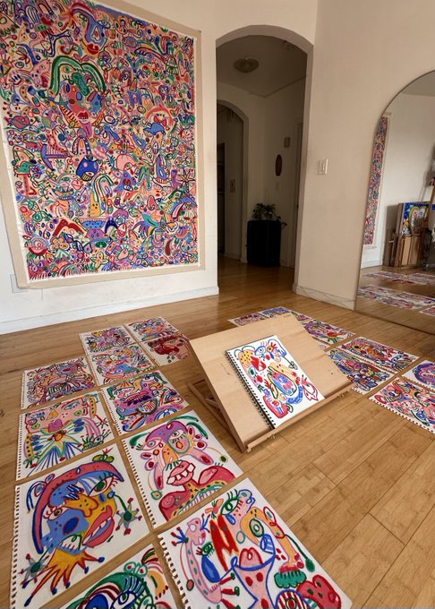An artist's studio displays a large, intricately detailed abstract painting on the wall alongside numerous smaller colorful artworks spread across the wooden floor and arranged in a sketchbook. The vibrant, psychedelic style of the artwork collections creates an inspiring creative atmosphere that perfectly complements the caption "These arts together look so nice!"