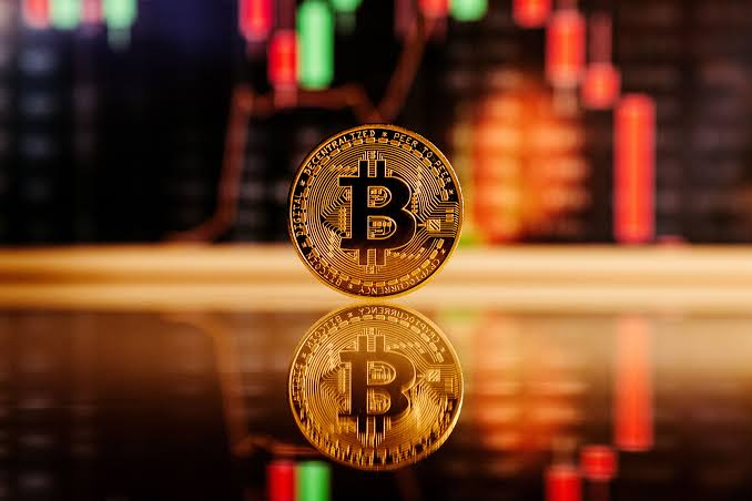 A physical Bitcoin token stands upright on a reflective surface, with its reflection visible below, set against a blurred backdrop of red and green candlestick trading charts. The coin's inscription reads "BITCOIN DIGITAL DECENTRALIZED PEER TO PEER" along its edge, visually reinforcing the article's focus on $BTC short-term holder profitability reaching historic highs of around $168 billion.