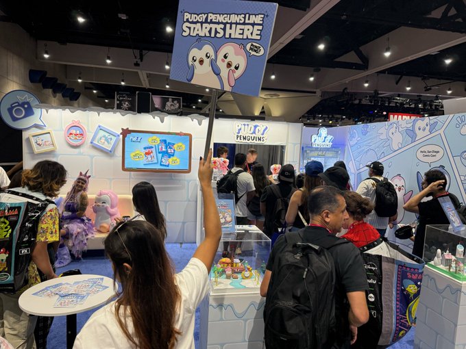 A person holding up a sign that reads 'PUGGY PENGUINS LINE STARTS HERE' at a busy convention booth for the Pudgy Penguins brand, with merchandise and attendees visible.