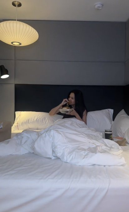 A woman sits up in bed eating from a container while surrounded by rumpled white sheets, illuminated by a modern hanging lamp in what appears to be a hotel room. The image captures the candid moment of her interrupted relaxation, humorously illustrating the post's frustration about missing a Stake release announcement just as she finally got comfortable.