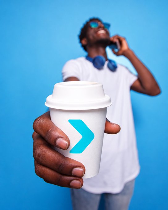 A young man with headphones around his neck holds out a coffee cup with a blue arrow logo toward the camera against a blue background, smiling while talking on a phone.