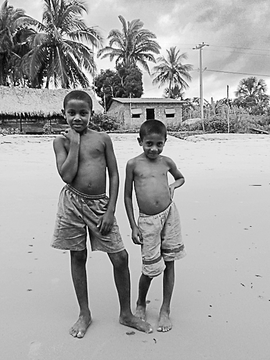 The image features two boys standing side by side on a beach, posing for a photo. They are both barefoot, enjoying their time at the shoreline. The background of the picture includes palm trees and houses, giving an impression of a coastal area with a tropical atmosphere. The image also contains text that reads "GN children! New listing ... Fishing Net Under Maintenance" and "Tezos".
