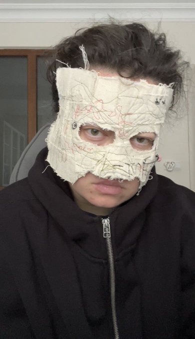 A selfie of a person with dark curly hair wearing a black hoodie and an unusual white fabric mask with stitched line patterns covering their face.