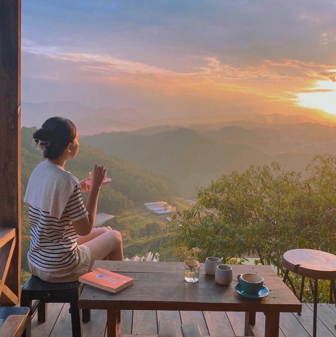 A woman sitting at a wooden table on a balcony, looking out at a scenic mountain landscape during a golden sunset.
