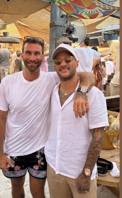 Two men pose smiling at the camera in a sunny outdoor setting, one wearing a white t-shirt and floral shorts while the other sports a white button-up, white cap, sunglasses, and a luxury watch, embracing the relaxed St. Tropez vibe mentioned in the post.