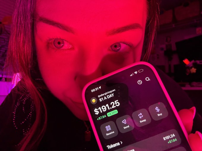 A woman bathed in red light holds up her phone displaying a crypto app showing a $1 daily purchase on @allergictoquack, with a current balance of $191.25 (+24.26%, +$7.84). The screen reflects her "day 225 of buying $1 worth of SOL until ATH" challenge, showing the accumulated value of her incremental Solana investments.