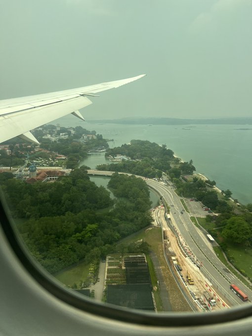 A traveler captures an aerial view from an airplane window, showing the wing of the plane over a coastal landscape with highways, greenery, and water below, likely approaching Singapore after a long 15-hour flight. The image reflects the anticipation of arriving in the city-state for an exciting meetup with the Hyperliquid (HL) community.