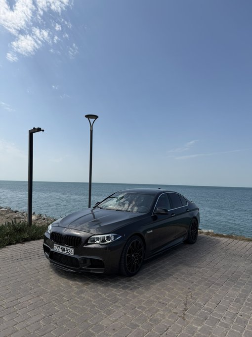 A dark-colored BMW sedan is parked on a stone-paved area beside the ocean under a blue sky, with license plate '777 AM 924 AZ'.
