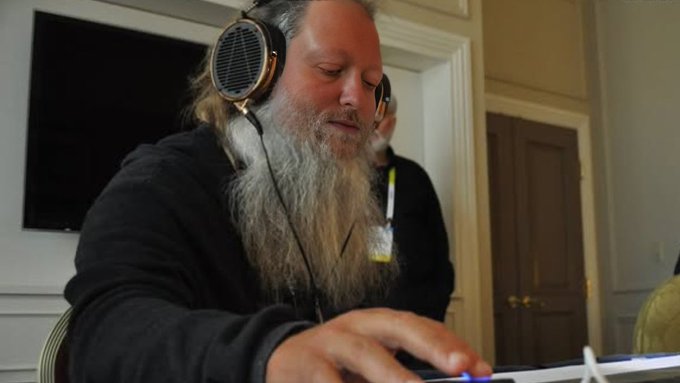 The image shows a man wearing headphones, sitting at a desk or table while using a computer mouse. The background features a TV screen mounted on the wall above him. The caption of the image reads "Focus on what is in the image and how it relates to the post." This suggests that the viewer should pay attention to the details within the image and consider its connection to the content displayed at the bottom of the photo, which appears to be a discussion about Dash or computer mice.