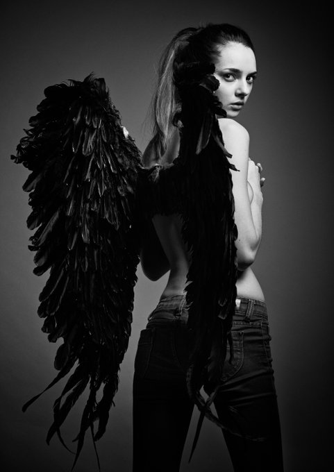 A black and white portrait features a woman with long hair tied back, wearing jeans and large black feathered wings, as she looks over her shoulder at the camera.