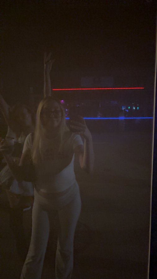 A dark photo showing a woman with long blonde hair and glasses posing while holding a phone, with a red neon light strip visible in the background.