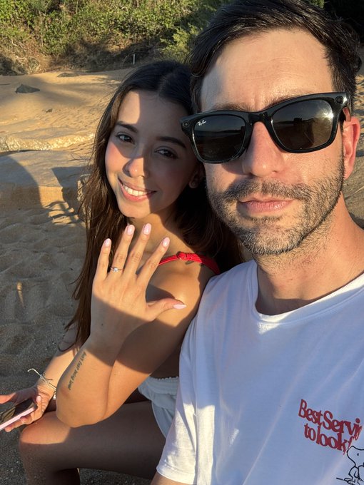 A selfie of a couple on a rocky beach, with the man wearing sunglasses and a white t-shirt, and the woman smiling and showing her hand as if displaying a ring.