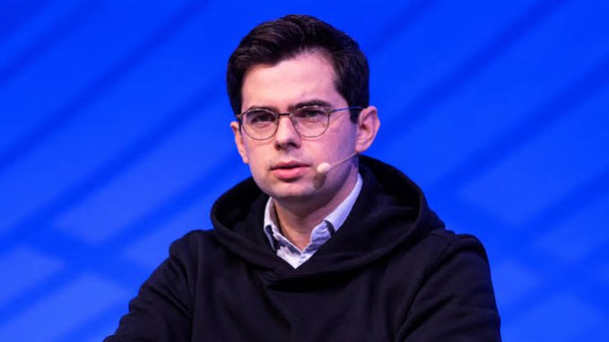 A man wearing glasses, a dark hoodie, and a headset microphone sits on a stage with a blue geometric backdrop.