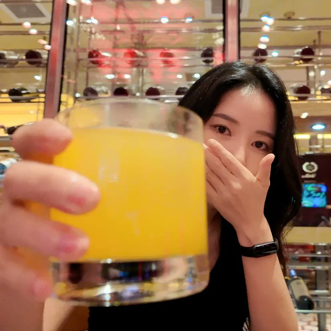 A young woman with long dark hair peeking over a glass of orange juice, covering her mouth with one hand, with a wine rack visible in the softly lit background.