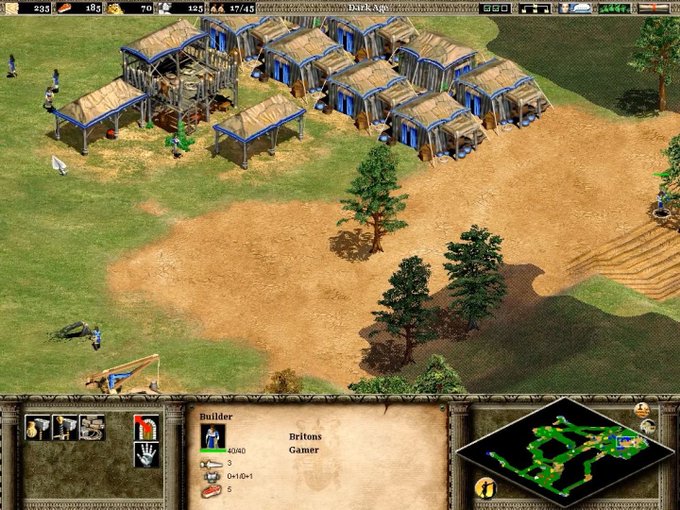 A screenshot from the strategy game Age of Empires showing a base with wooden buildings, trees, and a builder unit selected, labeled as a Britons player named 'Gamer'.