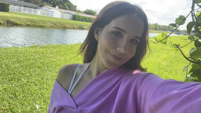 An outdoor selfie of a dark-haired woman wearing a purple off-the-shoulder top, posing on green grass next to a pond with houses in the background.