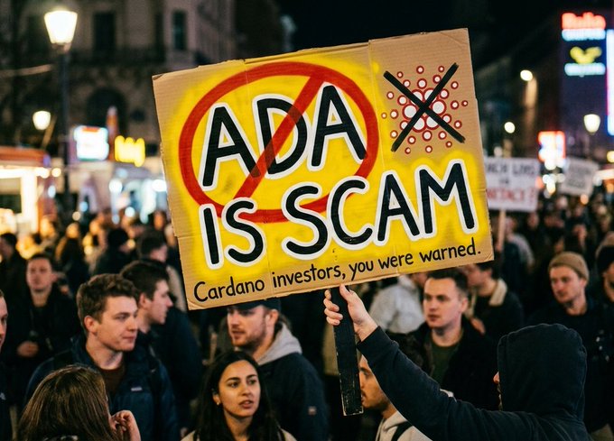 A protester at a nighttime gathering holds up a cardboard sign reading 'ADA IS SCAM' with a red prohibition symbol and 'Cardano investors, you were warned' written at the bottom.