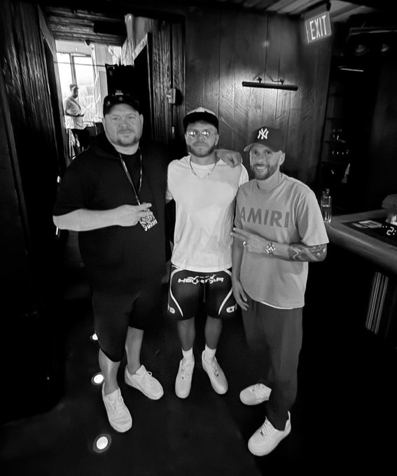 Three men pose together indoors; the man on the right wears a shirt reading 'AMIRI' and a Yankees cap.