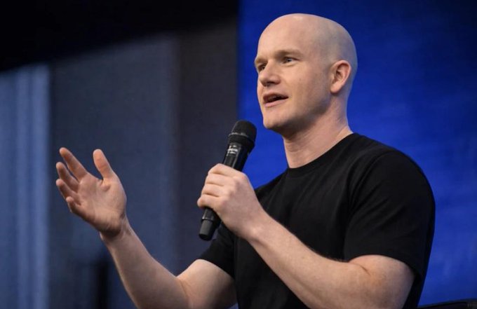 A bald man in a black t-shirt speaks into a microphone while gesturing with his hand against a blue backdrop. The image appears to depict Brian Armstrong, the Coinbase CEO mentioned in the post about XRP being usable as collateral for borrowing USDC.