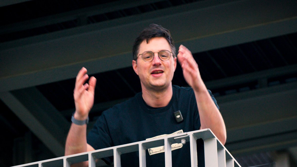The image features a man standing on a balcony with his hands raised up in the air. He appears to be wearing glasses, and there are two other people visible in the background who seem to be engaged in conversation or observing something happening below them. The scene suggests that they might be discussing the Jingle Miner Bitcoin hashrate calculator, which is an essential tool for miners to determine their mining power and potential rewards.