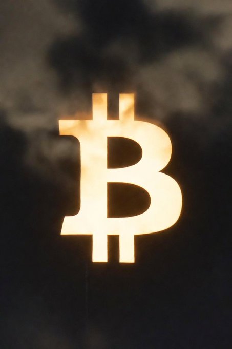 A glowing Bitcoin (₿) symbol illuminated against a dark, cloudy background.