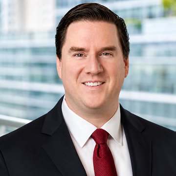 A professional headshot of a smiling man in a dark suit, white shirt, and red tie, photographed against a blurred office building backdrop.