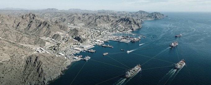 An aerial shot shows a coastal port with rugged mountains and several cargo ships navigating through a harbor, where overlaid lines appear to connect the vessels—visually illustrating how AIS and other tracking systems map out a ship's location and identity.