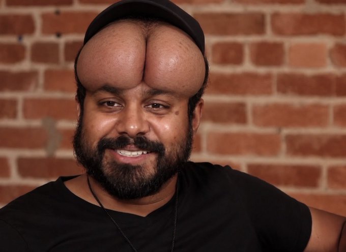 A smiling bearded man wearing a black cap and black t-shirt posing in front of a brick wall, with an obviously AI-edited distorted forehead and head shape.
