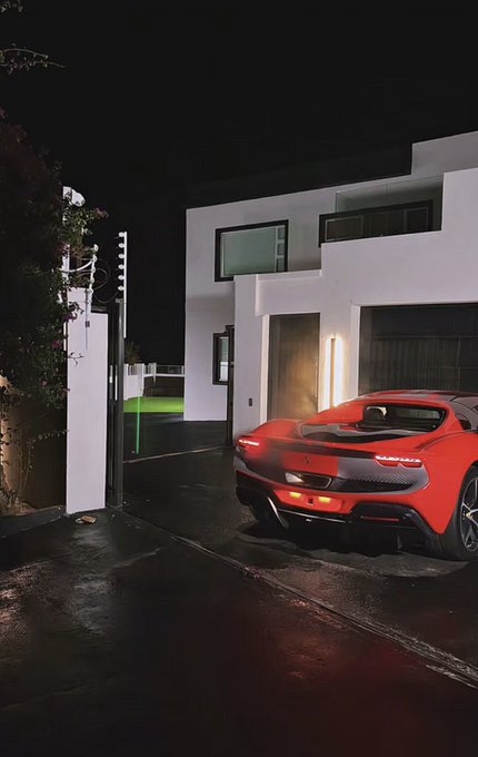 A red sports car parked in the driveway of a modern white minimalist house at night, with the wet pavement reflecting light.