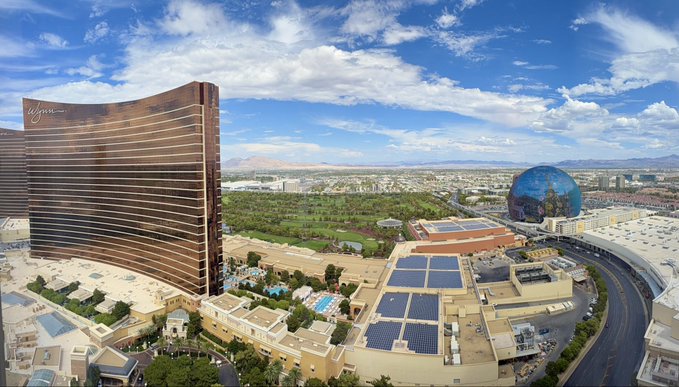 Aerial view of the Las Vegas Strip showing Wynn Las Vegas, Encore, and the MSG Sphere against a backdrop of mountains and partly cloudy sky.