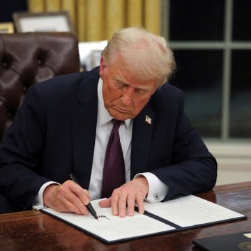 Donald Trump signing a document at the Resolute Desk in the Oval Office, wearing a dark suit and red tie.